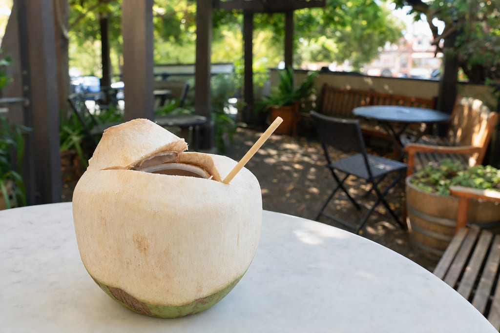 Fresh coconut with its top cut open and a paper straw standing in it, on a white marble table on the patio