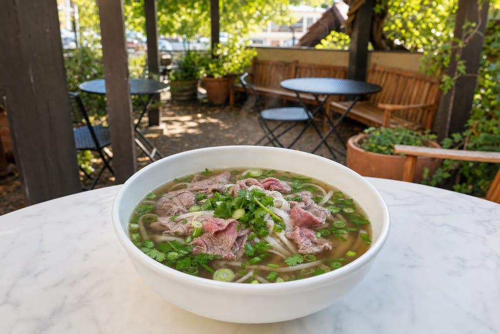Beef pho: thin slices of rare beef, rice noodles, green onion and cilantro in broth in a white bowl on the patio