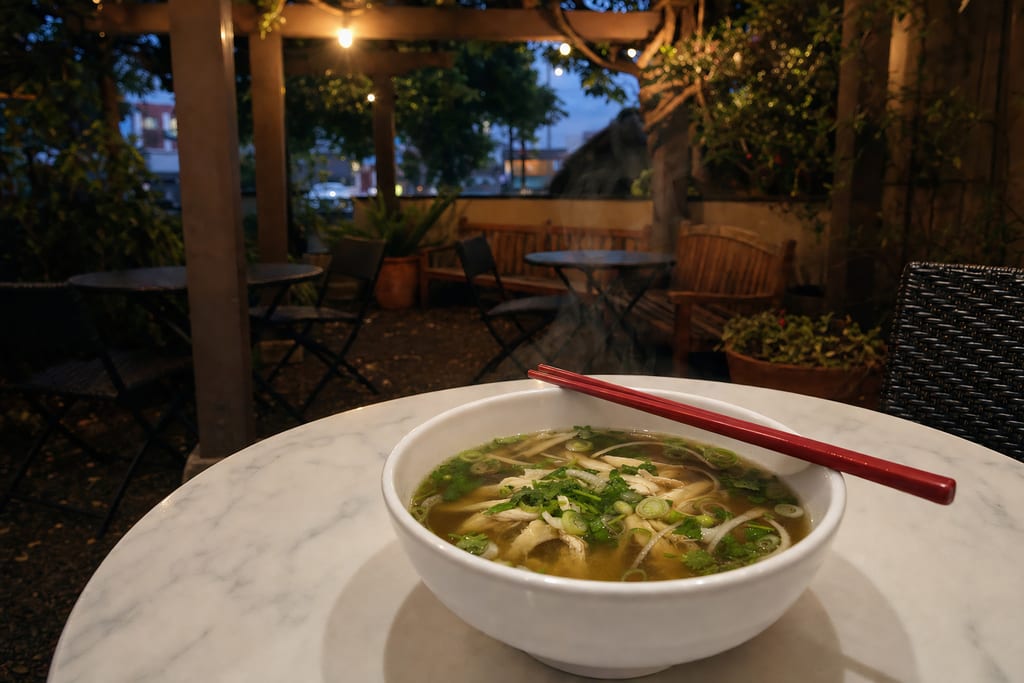 A steaming bowl of chicken pho with red chopsticks on the rim, on a marble patio table at dusk under string lights
