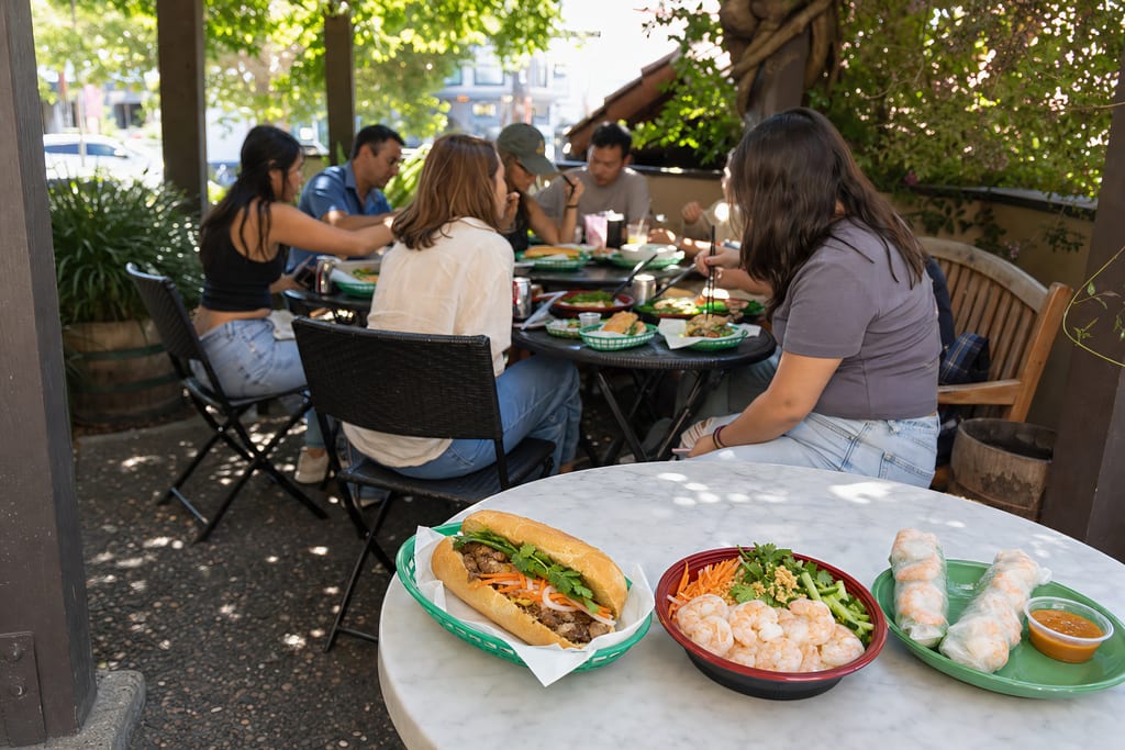 Friends sharing a meal at a patio bistro table, with a banh mi, a shrimp bowl and spring rolls on the marble table in front