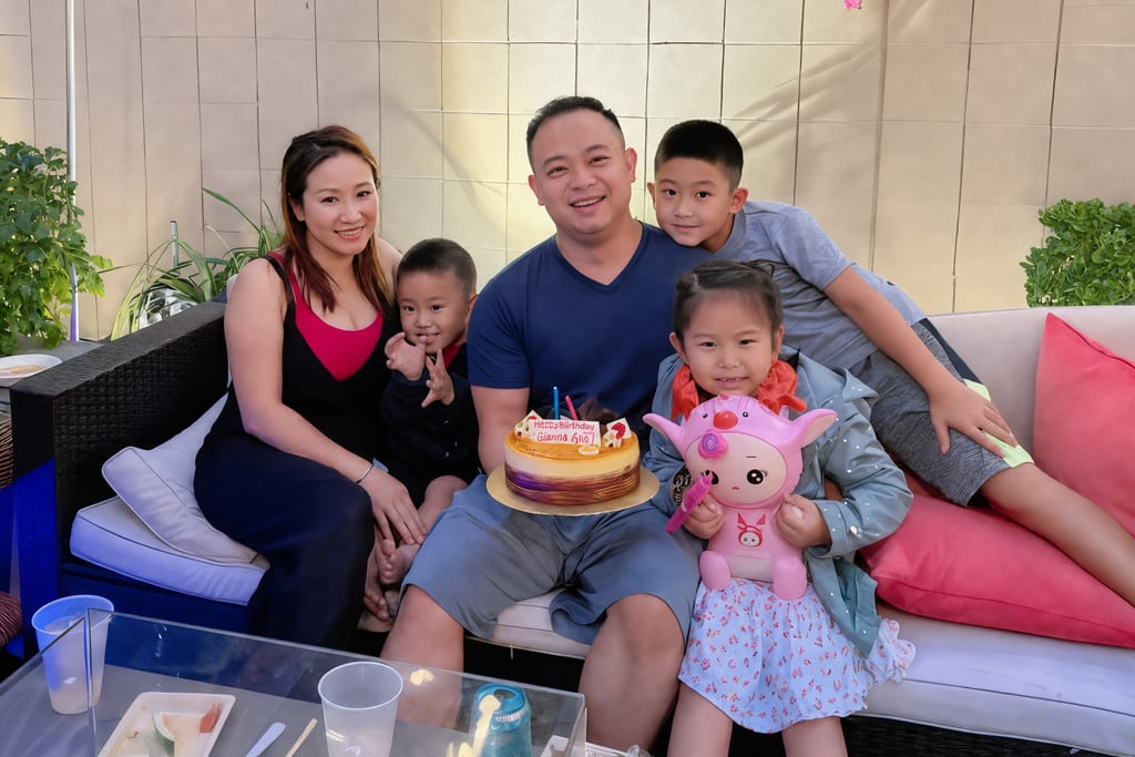 The Nguyen family on an outdoor sofa: a woman, a man holding a birthday cake, two boys and a girl holding a pink toy