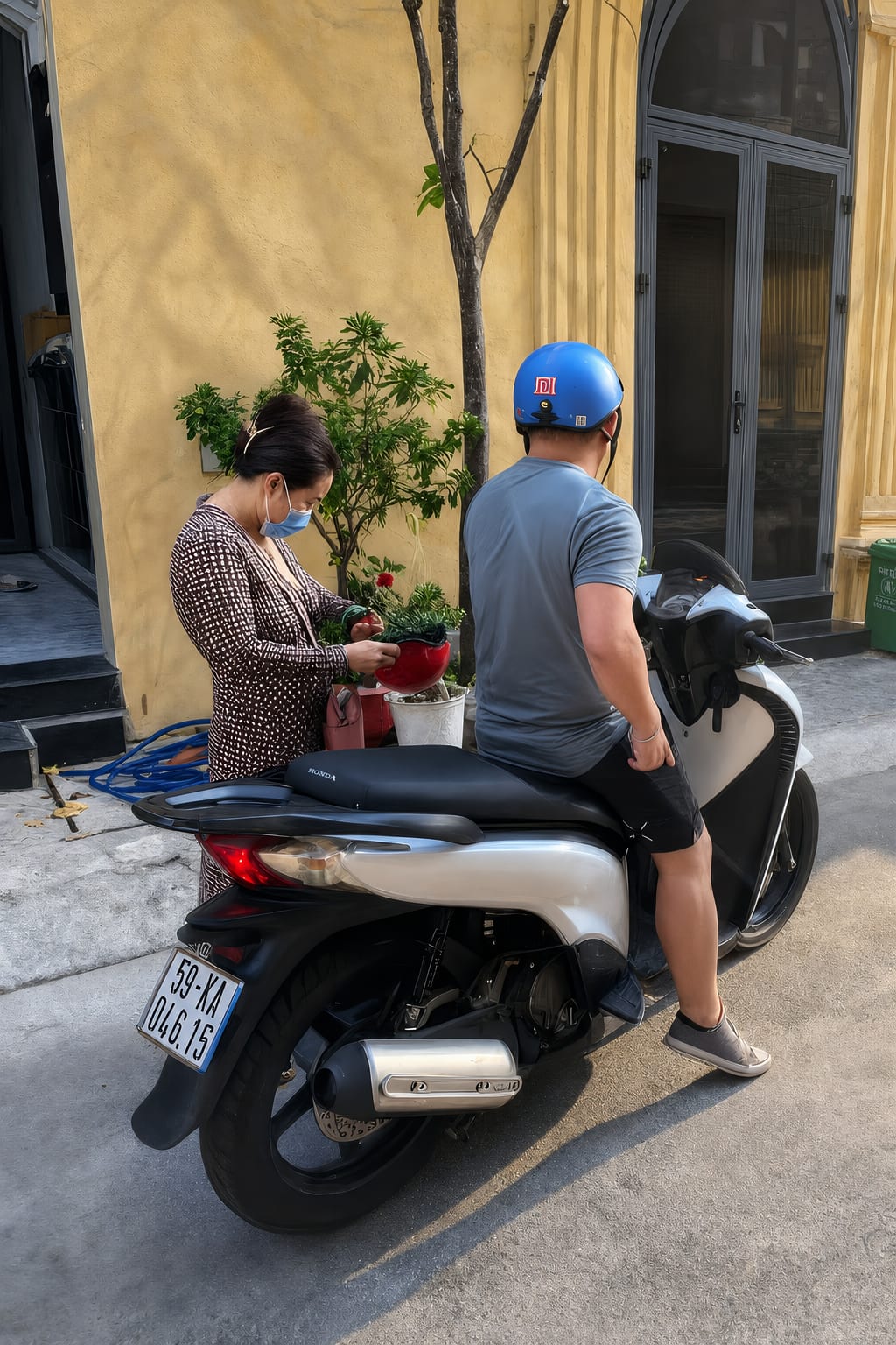 A man in a blue helmet on a scooter and a woman in a face mask tending a plant beside a yellow wall on a Saigon lane